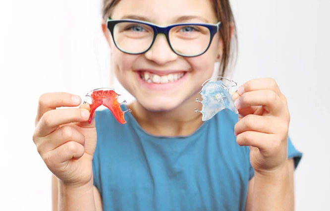 Lächelndes Mädchen mit blauer Brille und blauem Shirt, das zwei herausnehmbare Zahnspangen hält – eine rote und eine transparente.