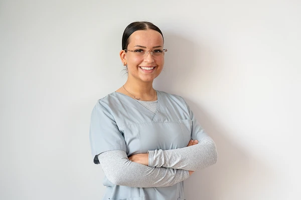 Lächelnde junge Mitarbeiterin mit Brille in grauer Praxiskleidung, die Arme verschränkt.
