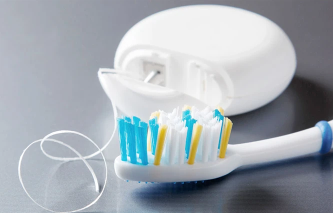 Nahaufnahme einer weißen Zahnbürste mit blauen und gelben Borsten neben einer Dose mit Zahnseide auf grauer Oberfläche.