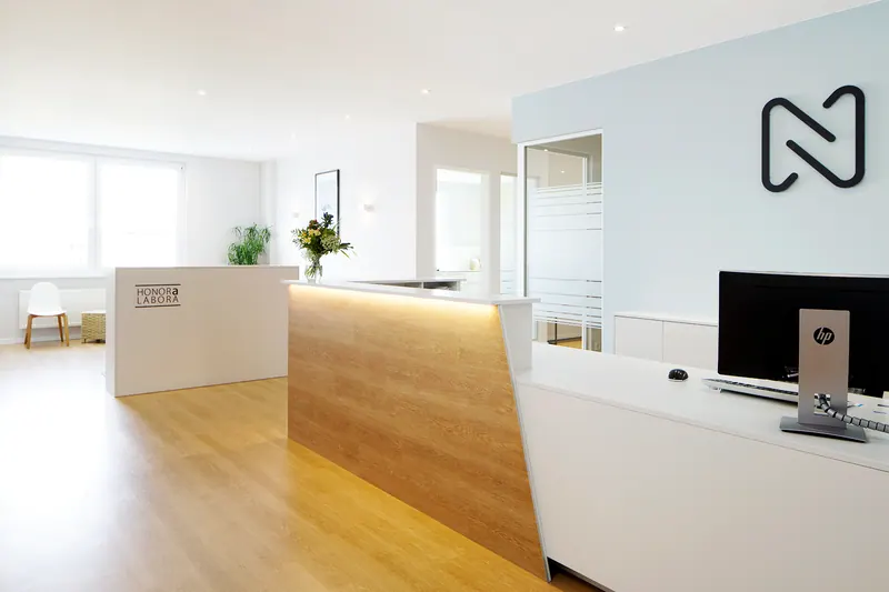 Moderner Empfangsbereich mit Holzboden, weißen Schreibtischen, einer Blumenvase, einem Computerbildschirm und viel Tageslicht durch große Fenster.