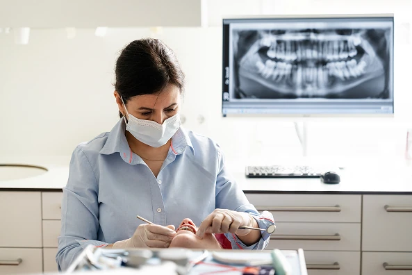 Behandlerin mit Mundschutz untersucht die Zähne einer Patientin, im Hintergrund ein Röntgenbild des Gebisses auf dem Monitor.