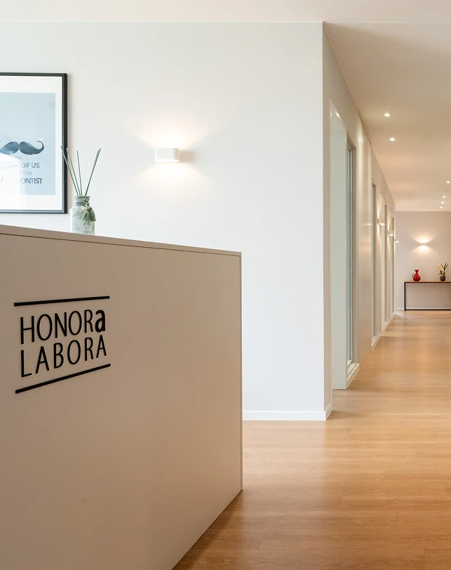 Moderne Praxisrezeption mit weißem Empfangstresen mit der Aufschrift „HONORA LABORA“, Holzboden sowie hellem Flur mit gerahmten Bildern und dekorativen Vasen.